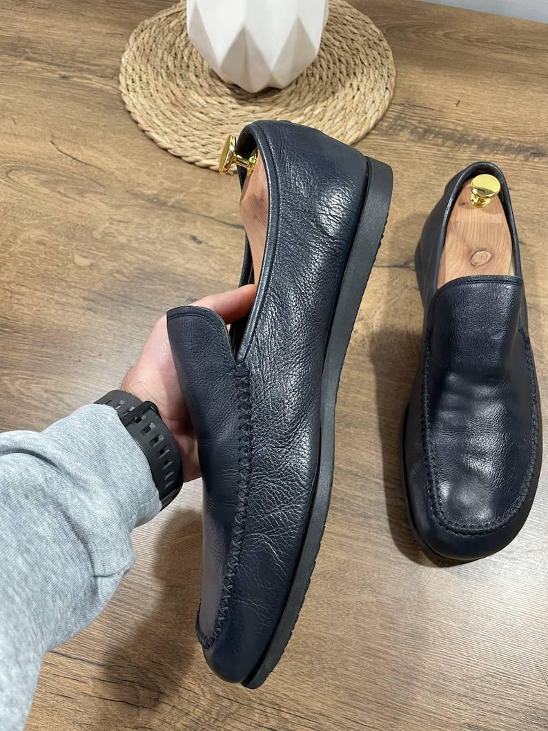 May include: Navy blue leather loafers with a rounded toe and stitched vamp. The shoes are displayed on a wooden surface. The loafers have a flat sole.