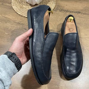 May include: Navy blue leather loafers with a rounded toe and a slip-on design. The shoes have a stitched seam detail around the edges and are shown with wooden shoe trees inside. The loafers are displayed on a wooden surface.