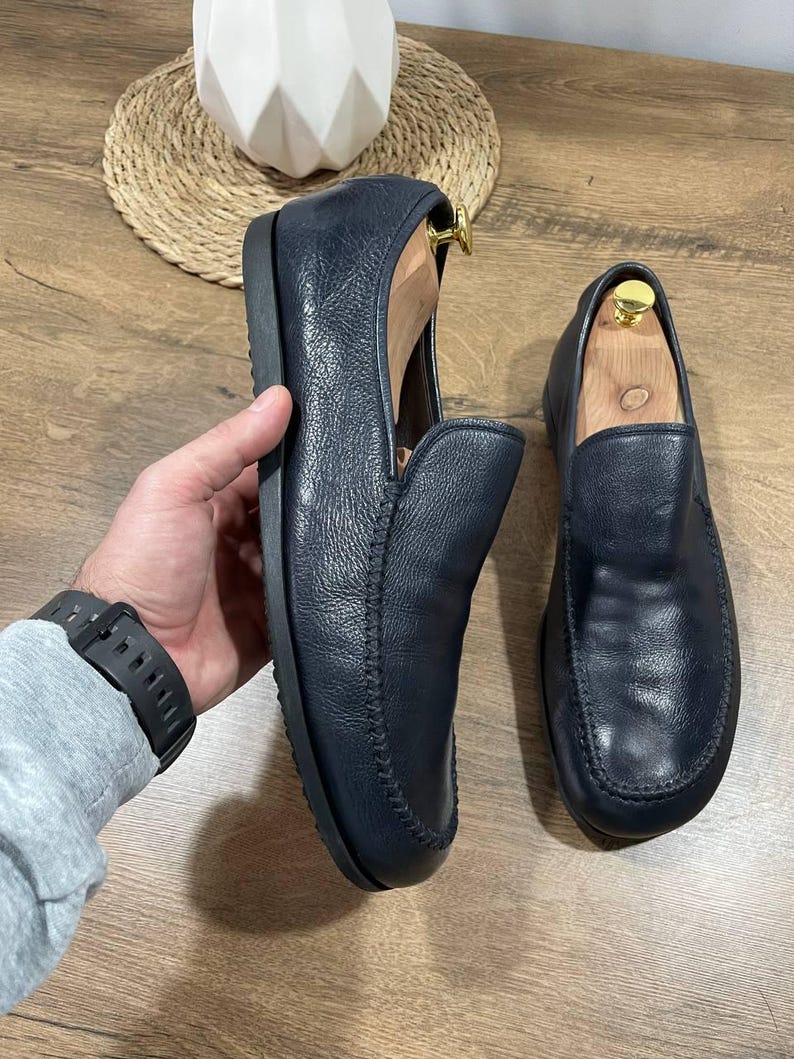 May include: Navy blue leather loafers with a rounded toe and a slip-on design. The shoes have a stitched seam detail around the edges and are shown with wooden shoe trees inside. The loafers are displayed on a wooden surface.