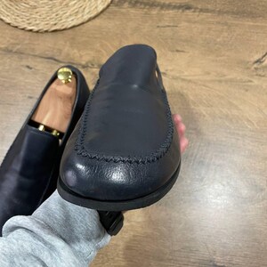 May include: Navy blue leather loafers, one in focus with a stitched seam detail. The other shoe, slightly blurred, has a wooden shoe tree inside. The shoes are displayed on a wooden surface. A classic footwear item.