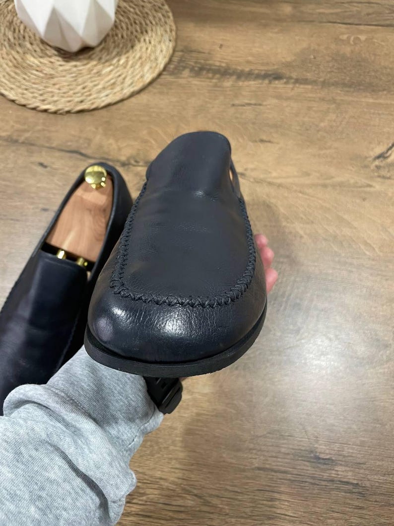 May include: Navy blue leather loafers, one in focus with a stitched seam detail. The other shoe, slightly blurred, has a wooden shoe tree inside. The shoes are displayed on a wooden surface. A classic footwear item.