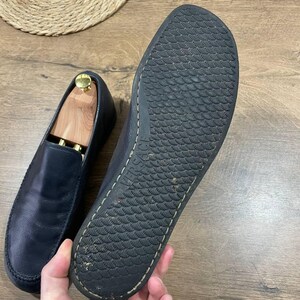 May include: Navy blue leather loafers. One shoe has a wooden shoe tree inside. The sole of the other shoe is visible, with the brand name "VERSACE" embossed. The soles have a textured pattern.