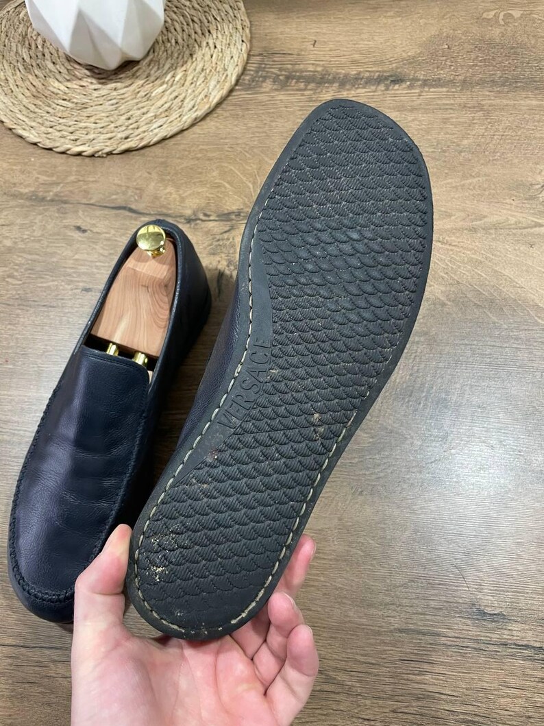 May include: Navy blue leather loafers. One shoe has a wooden shoe tree inside. The sole of the other shoe is visible, with the brand name "VERSACE" embossed. The soles have a textured pattern.