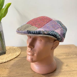 Może przedstawiać: Płaska czapka w stylu patchwork z czerwonymi, fioletowymi, zielonymi i beżowymi odcieniami. Czapka jest prezentowana na głowie manekina. Rondo czapki jest w kolorze jasnobeżowym. Czapka jest wykonana z teksturowanej tkaniny.