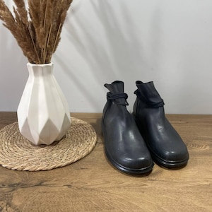 Puede incluir: Un par de botines de cuero negro con punta redondeada y suela gruesa. Las botas tienen un detalle fruncido en la parte superior y un lazo decorativo. Un jarrón blanco geométrico con plantas secas está a la izquierda.