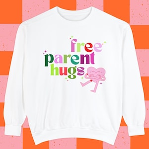 Puede incluir: Sudadera blanca de manga larga con el texto "free parent hugs" en letras de colores y un personaje de color rosa en forma de corazón con brazos y piernas que se abraza a sí mismo.