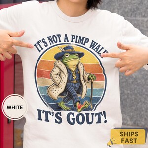 Könnte beinhalten: Ein weißes T-Shirt mit einer Grafik eines Frosches in einem blau gestreiften Anzug, Pelzmantel, Fedora und Goldkette. Der Frosch hält einen Gehstock und geht mit einem geschwollenen Fuß. Der Text lautet: IT'S NOT A PIMP WALK, IT'S GOUT! über einem Sonnenuntergang.