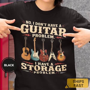 Puede incluir: Una camiseta negra de manga corta con un estampado gráfico de cinco guitarras diferentes y el texto: NO, I DON'T HAVE A GUITAR PROBLEM. I HAVE A STORAGE PROBLEM. El diseño incluye una guitarra acústica, una eléctrica azul claro, una roja de cuerpo hueco, una en forma de V y una eléctrica sunburst.