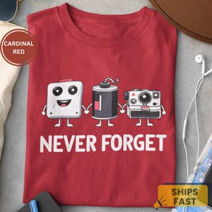 May include: Cardinal red t-shirt with a graphic of three cartoon camera-related items holding hands. The text "NEVER FORGET" is printed in white below the graphic. The shirt is a solid color.