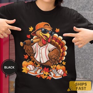 Puede incluir: Una camiseta negra con un gráfico de un pavo que lleva una gorra de béisbol naranja y gafas de sol, vestido con una camiseta de béisbol blanca. El pavo sostiene un bate, está junto a calabazas y pelotas, rodeado de hojas de otoño.