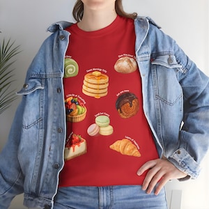 Puede incluir: Camiseta roja con ilustraciones de diversos productos de panadería y postres, como panqueques, cruasanes y macarons. La camiseta tiene texto que dice "Sweet Mornings Only", "A Little Bit of Everything", "Small but Fancy" y "Layers of Happiness".