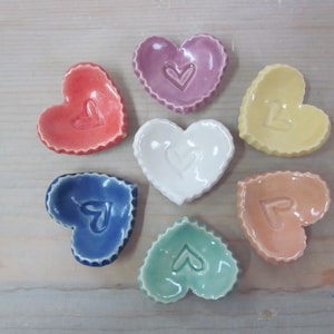 May include: Seven heart-shaped ceramic dishes in various colors, including pink, red, yellow, white, blue, orange, and green. Each dish has a smaller heart carved into the center.