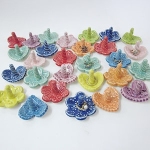 May include: A collection of colorful ceramic ring holders in various shapes, including hearts and flowers. Each holder features a raised, twisted knob for displaying rings.