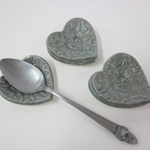 May include: Three gray ceramic heart-shaped spoon rests with a silver spoon resting on one of the rests.