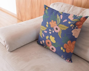May include: Square decorative pillow with a floral pattern. The pillow features a navy blue background with colorful flowers in shades of orange, pink, yellow, and green. The pillow is placed on a beige sofa.