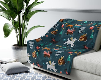 May include: A dark teal throw blanket featuring a repeating pattern of various dog breeds in festive attire, Christmas trees, and the text "MERRY WOOF MAS!". The blanket is draped over a white sofa, creating a cozy holiday theme.