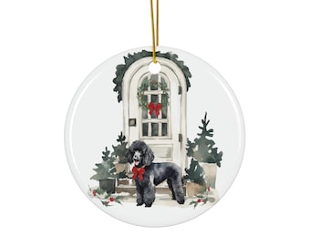 May include: A circular Christmas tree ornament depicting a black poodle with a red bow, positioned before a white door adorned with a wreath and red bow. The ornament has a gold hanging string and is decorated with Christmas trees.