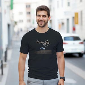 May include: Black t-shirt with the text "Moon Joy" in cursive script above an image of the moon's surface. The shirt is made of a soft material and is being worn by a person.