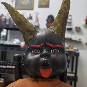 May include: A doll with black skin, two large textured gold horns, bright red eyebrows, and a red chin patch. The doll has light brown eyes, an open mouth expression, and wears a plain orange garment covering its torso.