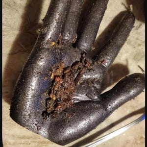 Puede incluir: Una escultura negra con forma de mano y acabado metálico texturizado. La palma está cubierta por un cúmulo de restos orgánicos oscuros, incluyendo pequeñas partículas marrones y rojizas. Una cuchara de metal plateado descansa en diagonal.