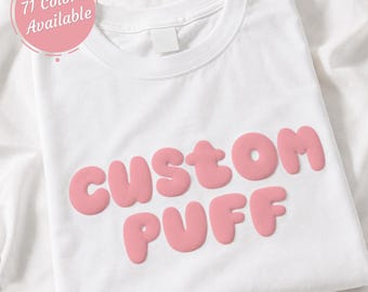 May include: A white crew neck t-shirt featuring the text CUSTOM PUFF in a raised, light pink, rounded font across the chest. A circular pink badge in the upper left corner contains the white text 71 Colors Available.
