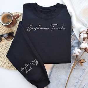 May include: A black sweatshirt with the words "Custom Text" in white script across the chest and on the left sleeve, accompanied by two hearts. The sweatshirt is displayed with a cup of coffee, sunglasses, and decorative cotton stems.