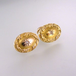 May include: A pair of gold cufflinks with a diamond in the center of each. The cufflinks are oval shaped and have a detailed design around the edge.