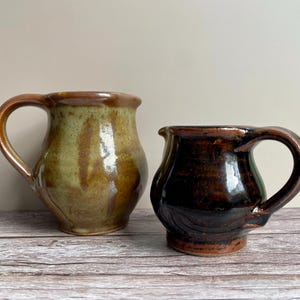 May include: Two handmade ceramic mugs. One is light brown with a green and brown swirl pattern. The other is dark brown with a glossy finish. Both have large handles and are on a wooden surface.