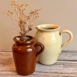 May include: Two ceramic vases. A small, glossy brown vase with a handle holds dried flowers. Beside it is a larger, off-white jug with a handle and a spout. Both are on a wooden surface.