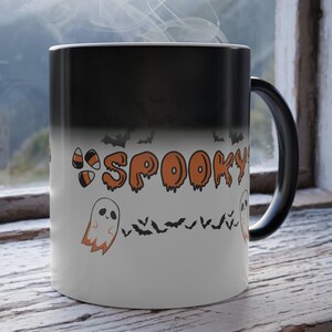 May include: A black and white color-changing mug with a spooky Halloween design. The mug features the word "SPOOKY" in orange and black dripping letters, a ghost, and bats. The mug is sitting on a wooden surface.