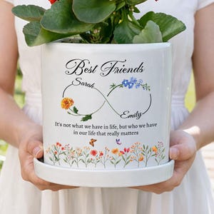 May include: A white ceramic flower pot with a floral design and the words "Best Friends" and the names "Sarah" and "Emily". The pot features an infinity symbol and the quote "It's not what we have in life, but who we have in our life that really matters."