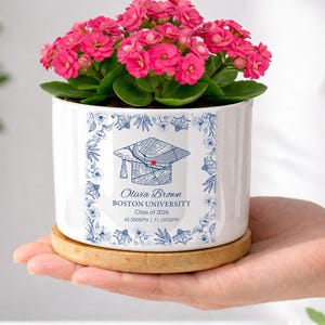 May include: A white ceramic planter with a graduation cap design and the text "Olivia Brown, Boston University, Class of 2026". The planter holds a flowering plant with vibrant pink blooms and sits on a wooden coaster.