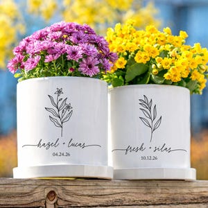 May include: Two white ceramic flower pots with floral designs and personalized text. One pot holds purple flowers, the other yellow. The text reads "hazel + lucas 04.24.26" and "fresh + silas 10.12.26".