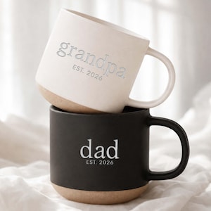 May include: Two ceramic mugs, one stacked on top of the other. The top mug is off-white with "grandpa" and "EST. 2026" printed on it. The bottom mug is black with "dad" and "EST. 2026" printed on it. Both mugs have a tan base and a curved handle.