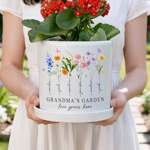 May include: White ceramic plant pot with a floral design and the text "GRANDMA'S GARDEN love grows here." The pot features colourful flowers and the names "amelia, josh, caroline, jennifer, madison, jack." A red flowering plant sits inside.