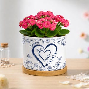 May include: A white ceramic flower pot with a heart design and the words "Grandma" and "Mom" in navy blue. The pot is filled with a flowering plant with bright pink blooms. The pot sits on a wooden saucer.