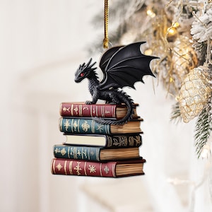 May include: A black dragon ornament with red eyes and large wings, perched atop a stack of books. The books have red, green, and tan covers. The ornament hangs from a gold string, suitable for a Christmas tree.