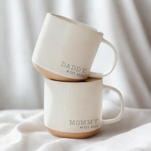 May include: Two stacked white ceramic mugs with a speckled finish and a natural brown base. The top mug reads "DADDY EST. 2026" and the bottom mug reads "MOMMY EST. 2026". Both mugs have a curved handle.