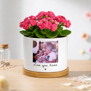 May include: A white ceramic planter on a circular wooden base containing a vibrant Kalanchoe plant with bright pink flowers. The front of the planter features a printed photograph of an adult and a child with the text Love you Nana below it.
