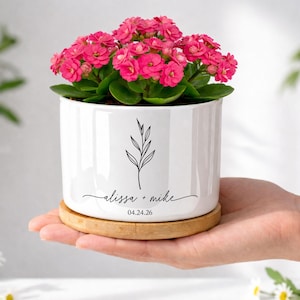 May include: A white cylindrical ceramic planter containing a vibrant pink Kalanchoe plant. The planter features a black line drawing of a leafy stem, the text "alissa + mike", and the date "04.24.26" printed on the front, resting on a bamboo base.