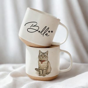 May include: Two stacked white ceramic mugs. The top mug has the name "Bella" in black script with paw prints. The bottom mug features a detailed illustration of a gray tabby cat wearing a red collar. Both mugs have a tan base and white handle.