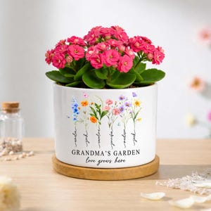 May include: A white ceramic cylindrical planter sitting on a circular wooden base, containing a vibrant pink Kalanchoe plant. The planter features a colourful floral illustration with the text "GRANDMA'S GARDEN love grows here" and names.