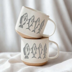 May include: Two stacked ceramic mugs with a cream-colored exterior and a natural brown base. Each mug features a series of gray fish illustrations. The mugs have a simple, rounded handle.