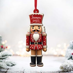 May include: A red and white Christmas ornament shaped like a nutcracker soldier. The ornament has a red hat with the name "James" and the year "2025" printed on it. The nutcracker is wearing a red uniform with black boots and a white beard. A red ribbon is attached for hanging.