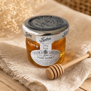 May include: A small jar of Tiptree Pure Honey with a silver lid and a wooden honey dipper. The jar contains 1 oz of honey, and the label reads "Wilkin & Sons Ltd, Tiptree Essex England". The honey dipper is made of light-colored wood.
