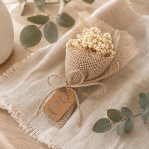 May include: A small bouquet of dried, cream-colored flowers wrapped in burlap. The bouquet is tied with twine and a small tag that reads "Thank You". The arrangement sits on a beige linen surface, with eucalyptus leaves in the background.