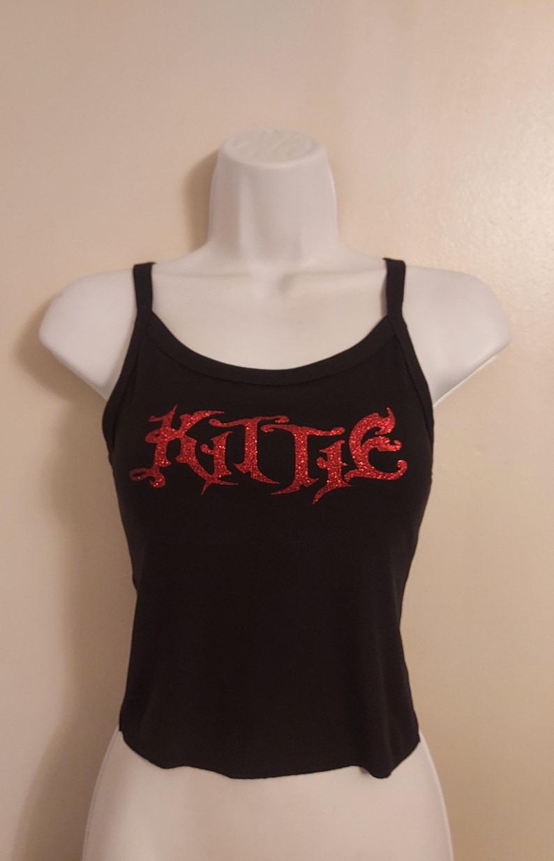 Puede incluir: Top negro de tirantes con letras rojas de purpurina que dicen "KUTIE".