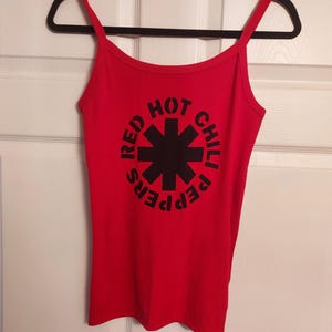 Puede incluir: Camiseta de tirantes roja de Red Hot Chili Peppers con un logotipo negro con una estrella y el nombre de la banda.