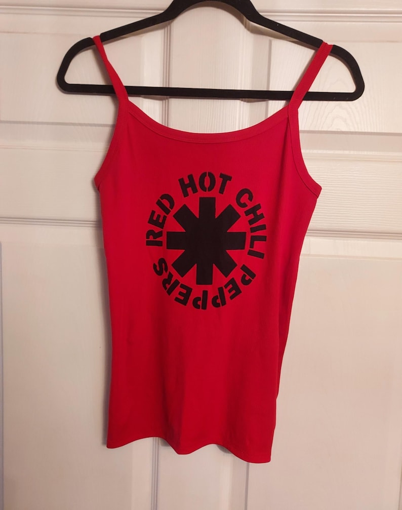 Puede incluir: Camiseta de tirantes roja de Red Hot Chili Peppers con un logotipo negro con una estrella y el nombre de la banda.