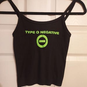 Może przedstawiać: Czarny top na ramiączkach z napisem "TYPE O NEGATIVE" w jaskrawozielonym kolorze, wraz z zielonym symbolem koła i linii poziomej. Top ma cienkie ramiączka i wisi na czarnym wieszaku.